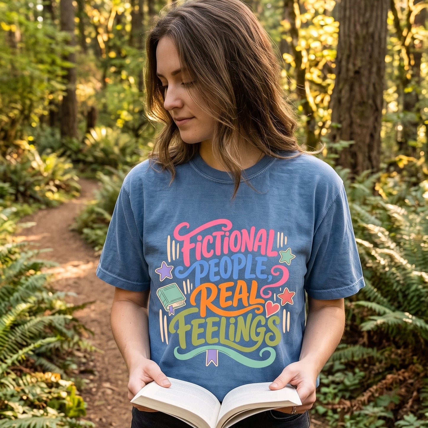 Woman wearing a Blue Jean Fictional People, Real Feelings book lover T-shirt while reading outdoors in a woodland setting.