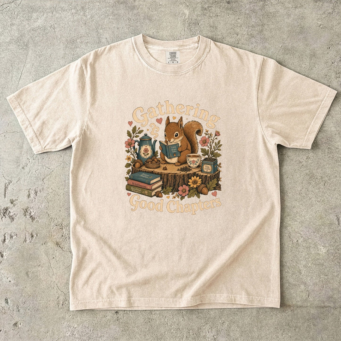 Front view of the Gathering Good Chapters Book Lover T-shirt on a Comfort Colors® Ivory shirt featuring a cozy woodland reading scene with a squirrel, books, tea and botanical details.