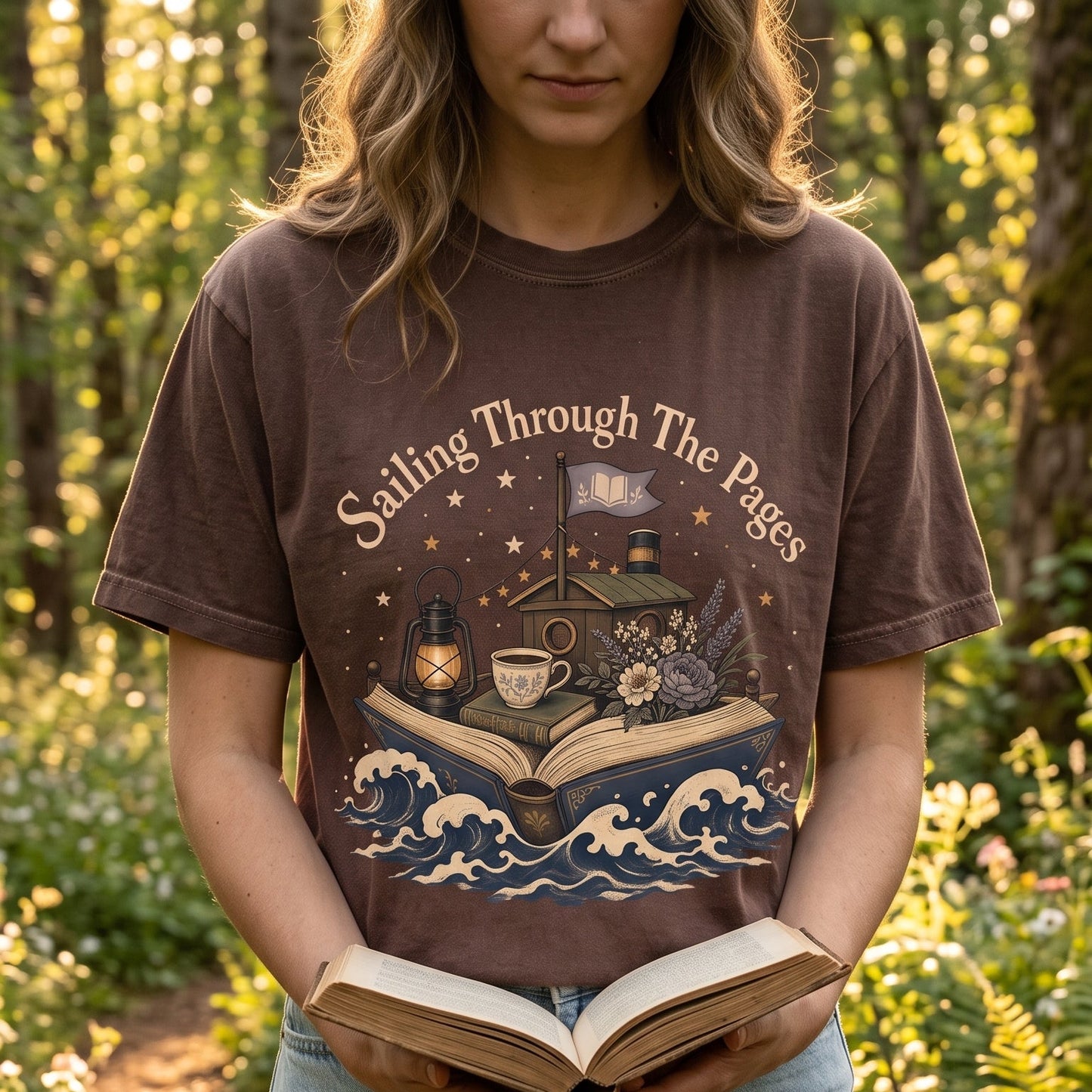 Woman wearing the Sailing Through the Pages Comfort Colors® Book Lover T-shirt while reading outdoors, featuring a vintage illustration of an open book sailing through ocean waves with a cozy cottage and lantern.