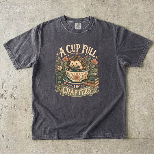 A Cup Full of Chapters Book Lover Shirt printed on a Comfort Colors 1717 garment-dyed T-shirt in Pepper featuring a cosy hedgehog reading inside a teacup with books and flowers.