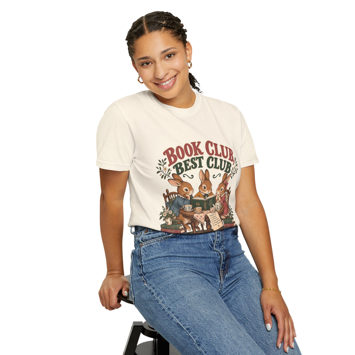 A woman wearing a ivory-colored graphic t-shirt with a 'Book Club Best Club' illustration of animals reading, paired with blue jeans, posing against a solid white background.