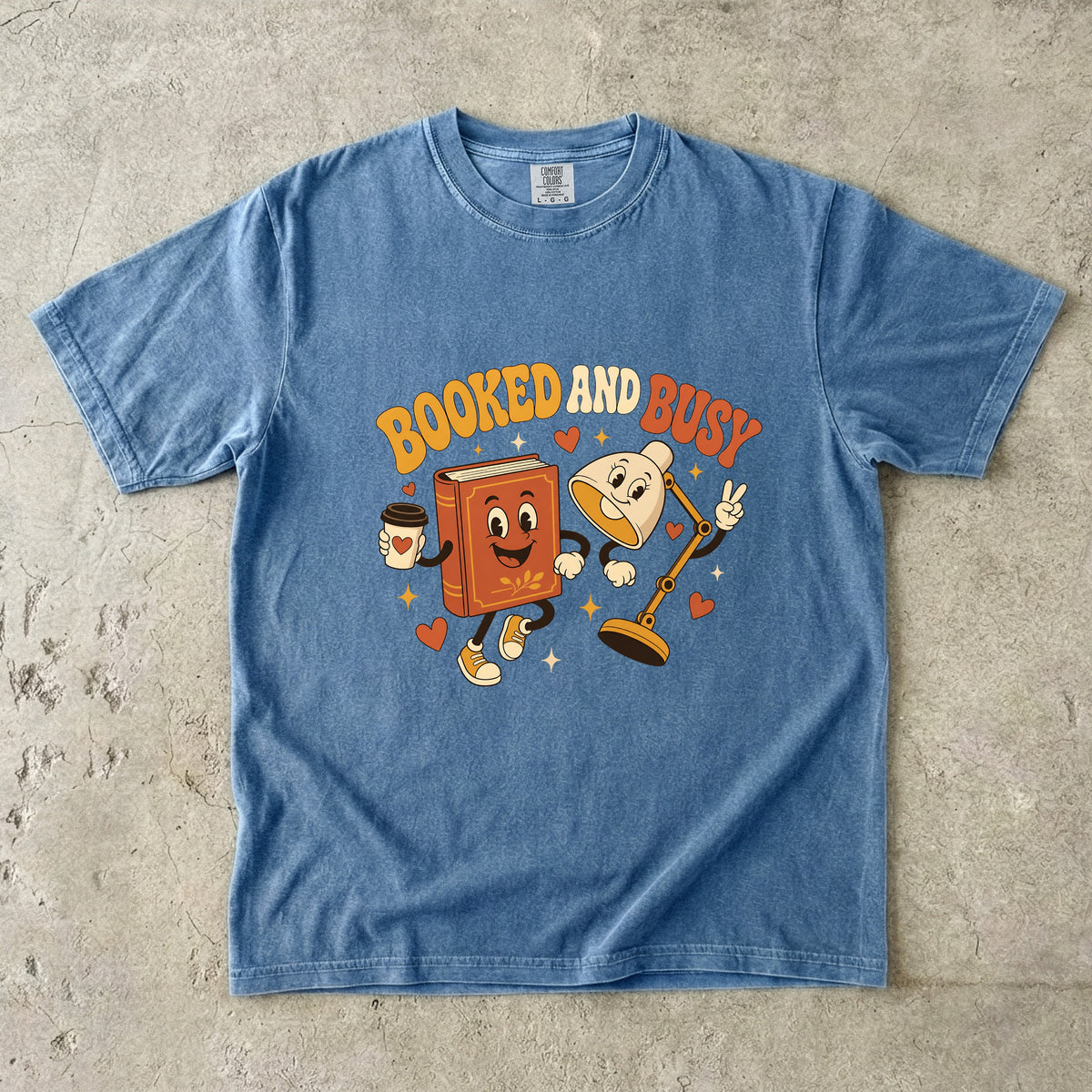 Blue Jean book lover T-shirt featuring Booked and Busy with a smiling book character, animated reading lamp, coffee cup, hearts and stars.