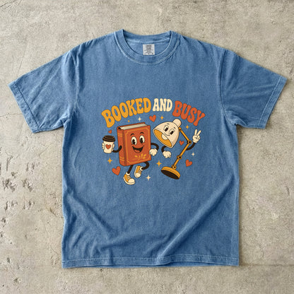 Blue Jean book lover T-shirt featuring Booked and Busy with a smiling book character, animated reading lamp, coffee cup, hearts and stars.