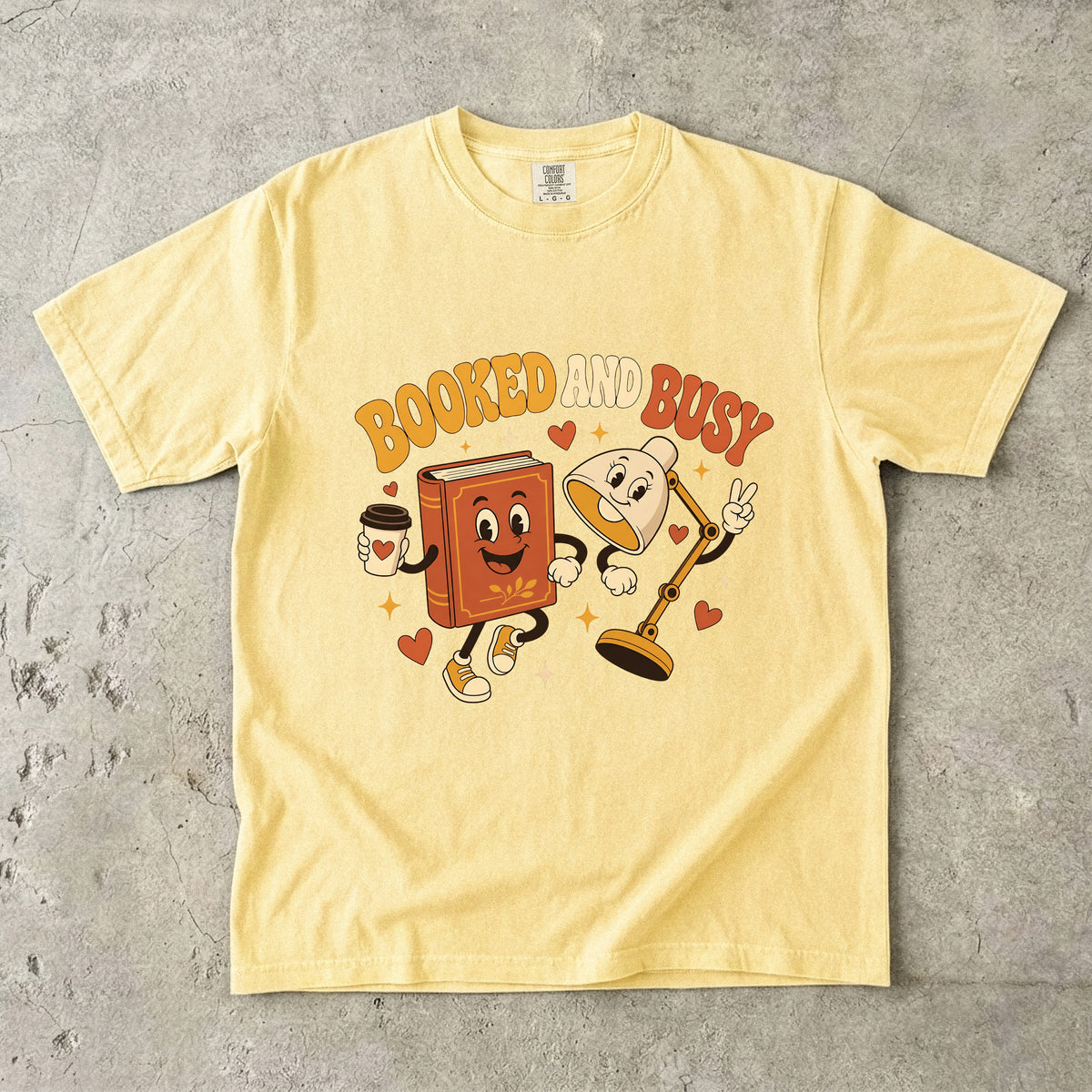 Butter book lover T-shirt featuring Booked and Busy with a smiling book character, animated reading lamp, coffee cup, hearts and stars.