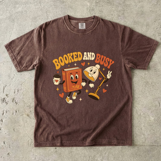 Espresso book lover T-shirt featuring Booked and Busy with a smiling book character, animated reading lamp, coffee cup, hearts and stars.