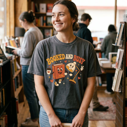 Booked and Busy book lover T-shirt featuring a playful smiling book, animated reading lamp, coffee cup, hearts and retro details.