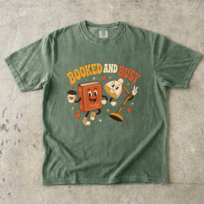 Moss book lover T-shirt featuring Booked and Busy with a smiling book character, animated reading lamp, coffee cup, hearts and stars.