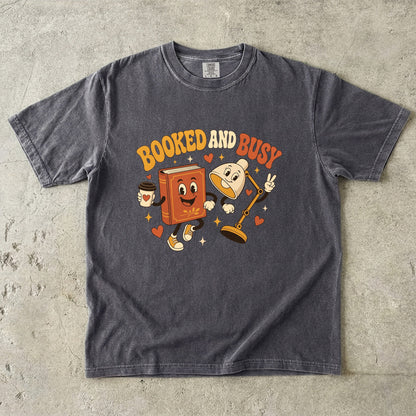 Pepper book lover T-shirt featuring Booked and Busy with a smiling book character, animated reading lamp, coffee cup, hearts and stars.