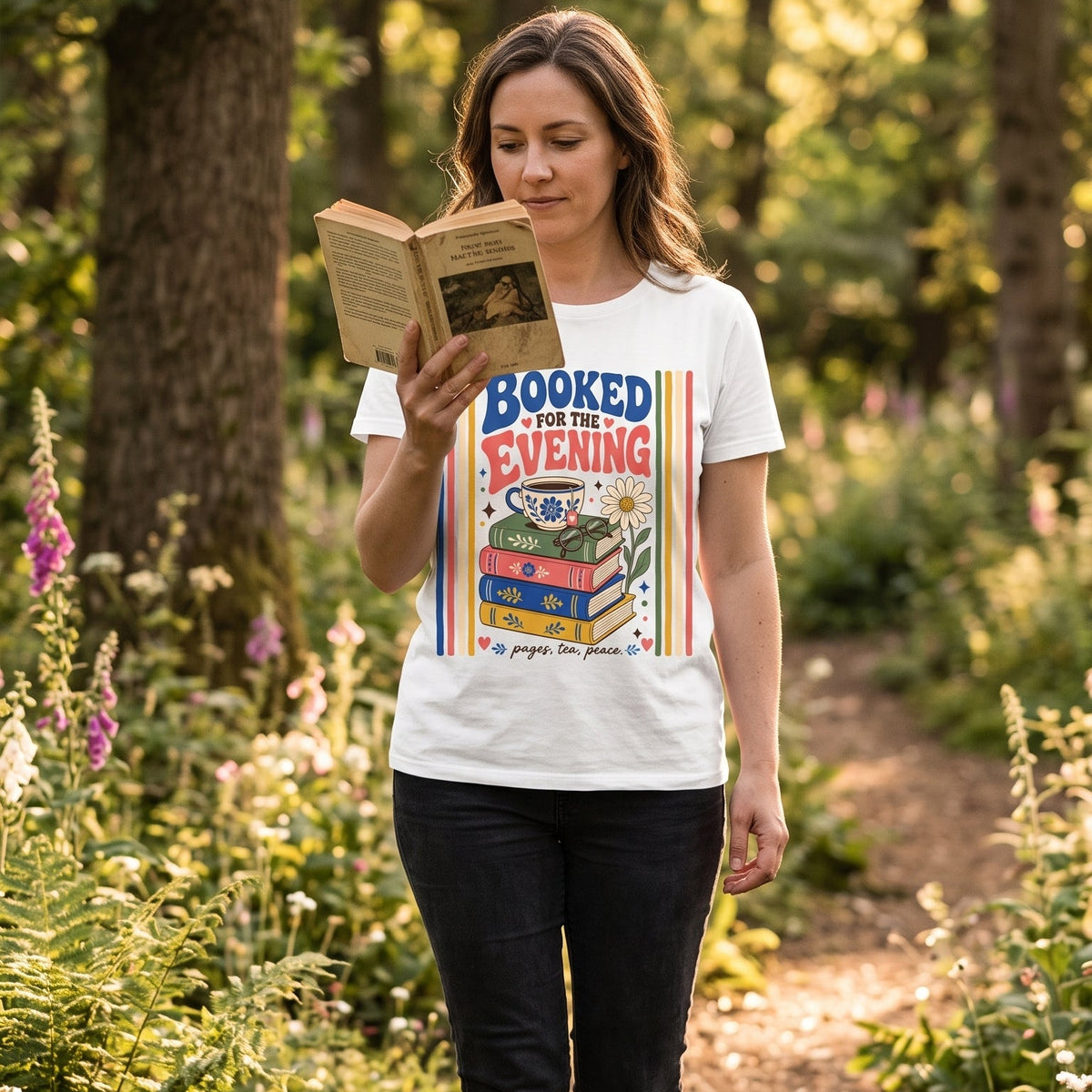Woman wearing the Booked for the Evening book lover T-shirt while reading outdoors, featuring stacked books, tea, glasses and a daisy.