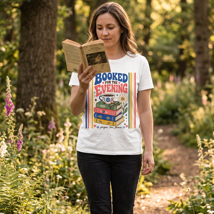 Woman wearing the Booked for the Evening book lover T-shirt while reading outdoors, featuring stacked books, tea, glasses and a daisy.