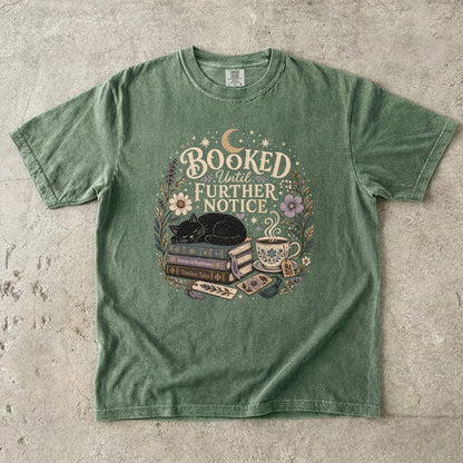 Booked Until Further Notice cat reading T-shirt with sleeping black cat, stacked books, teacup and flowers on Moss.