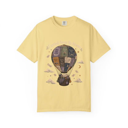Carried Away by Stories book lover T-shirt in Butter featuring a hot air balloon made from books with floating pages and clouds