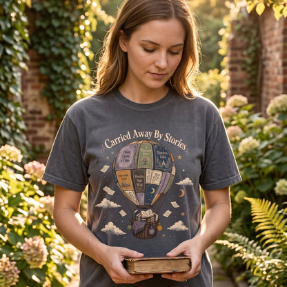Carried Away by Stories book lover T-shirt featuring a whimsical hot air balloon made from books surrounded by floating pages, clouds and stars