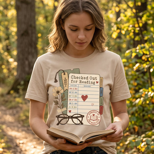 Woman wearing a Checked Out for Reading book lover T-shirt while reading a book outdoors.