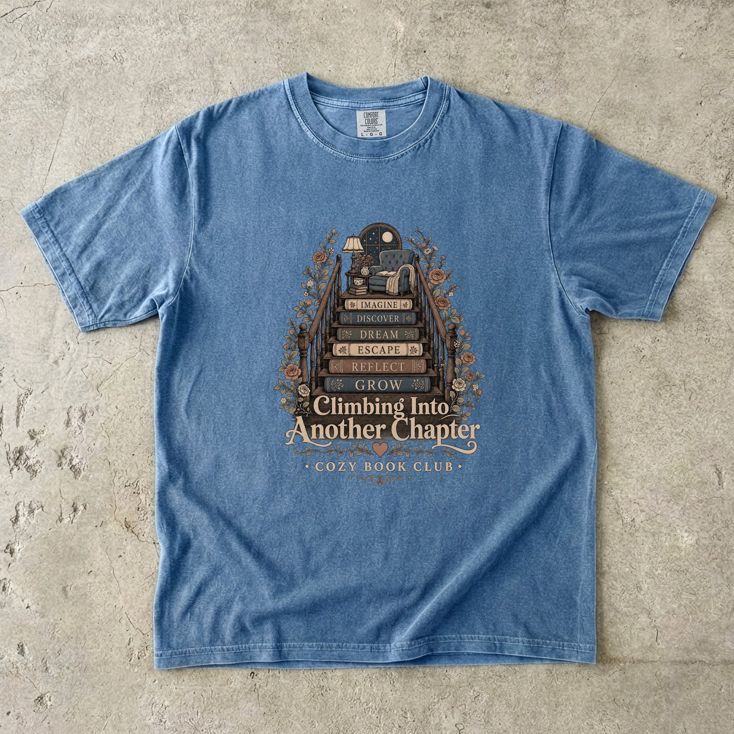 Front view of a Blue Jean Comfort Colors® book lover T-shirt featuring the Climbing Into Another Chapter design with stacked books forming stairs to a cozy reading chair beneath a moonlit window.