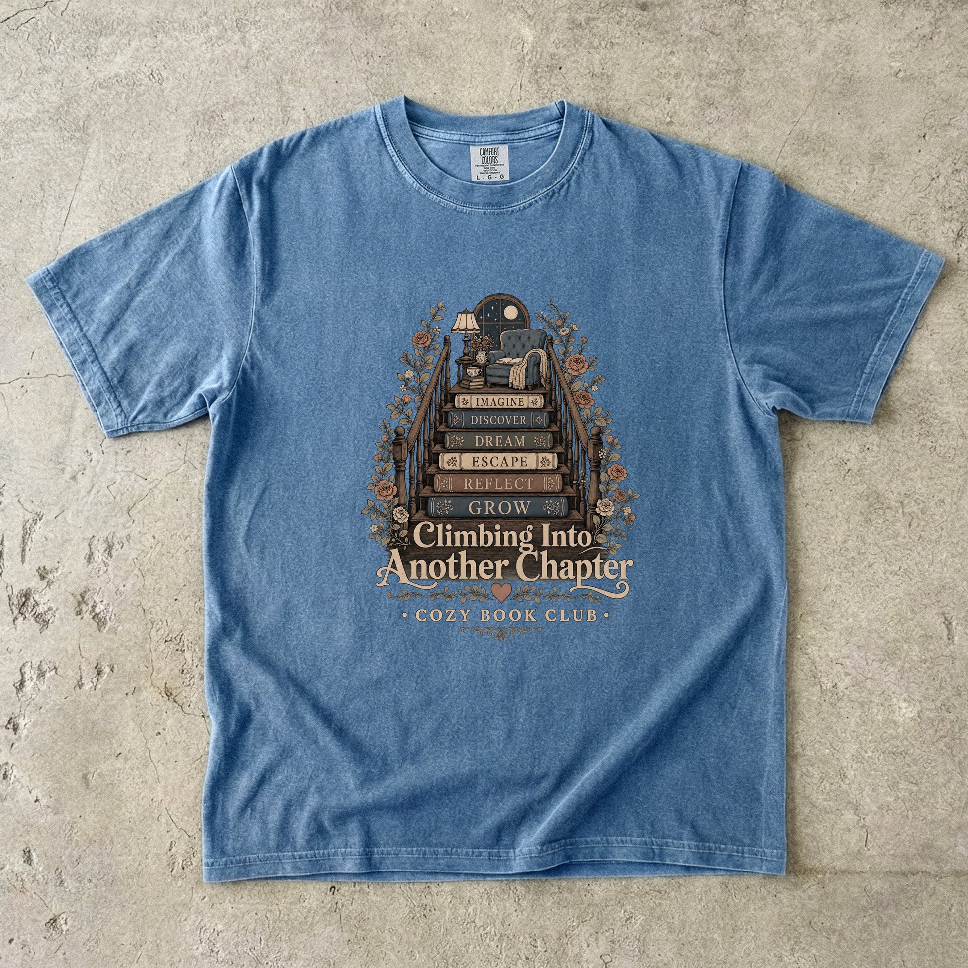 Front view of a Blue Jean Comfort Colors® book lover T-shirt featuring the Climbing Into Another Chapter design with stacked books forming stairs to a cozy reading chair beneath a moonlit window.