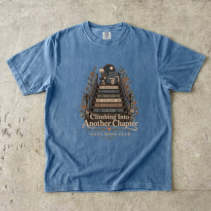 Front view of a Blue Jean Comfort Colors® book lover T-shirt featuring the Climbing Into Another Chapter design with stacked books forming stairs to a cozy reading chair beneath a moonlit window.