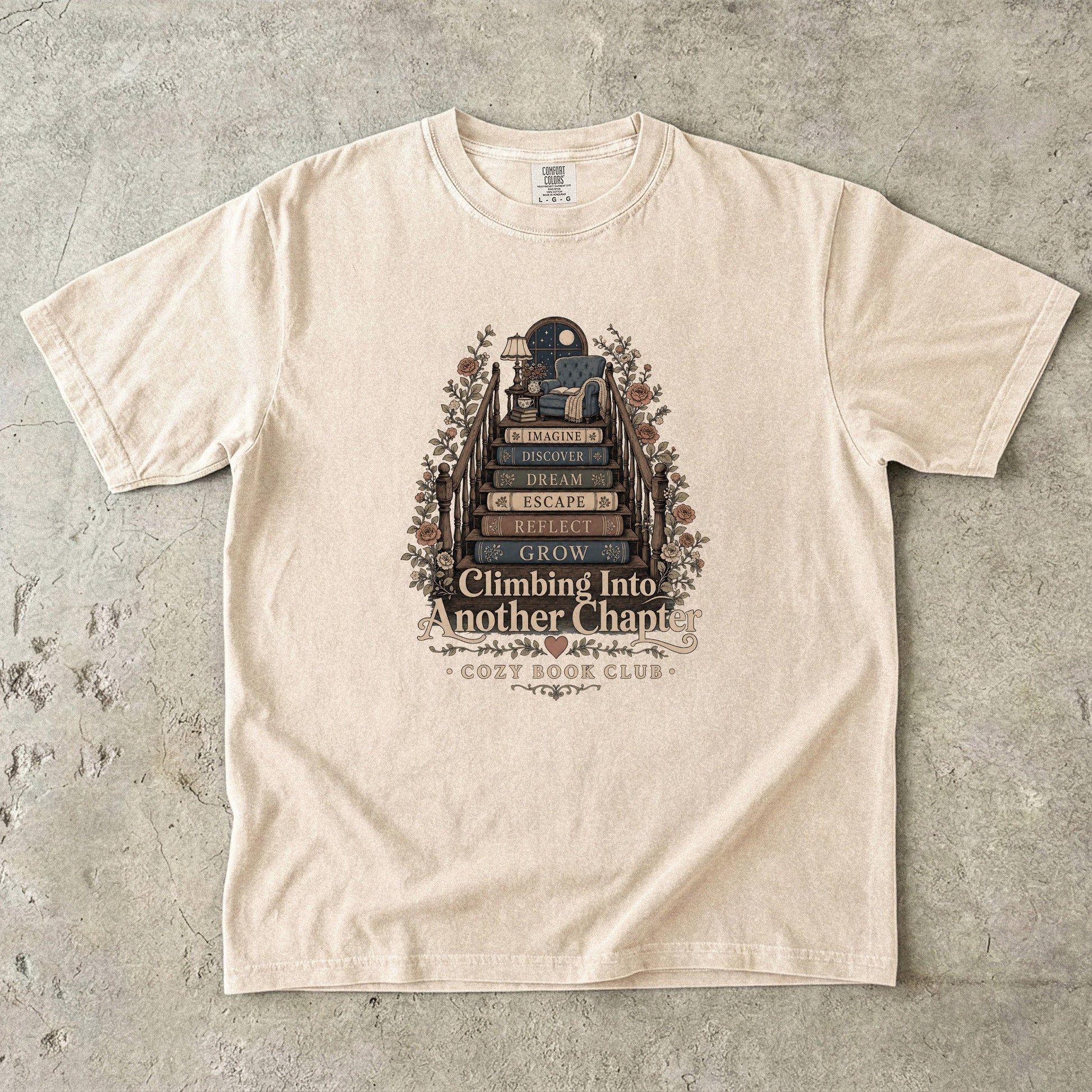 Front view of an Ivory Comfort Colors® book lover T-shirt featuring the Climbing Into Another Chapter design with stacked books, floral details and a moonlit reading retreat.