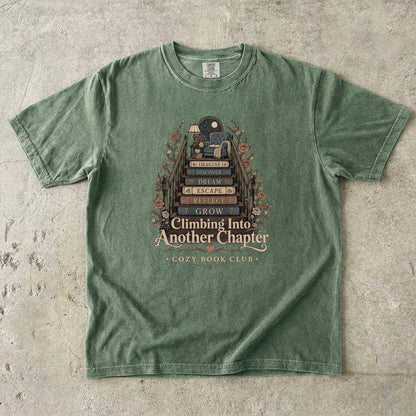 Front view of a Moss Comfort Colors® book lover T-shirt featuring the Climbing Into Another Chapter illustration with book stairs, cozy chair, botanical accents and literary artwork.