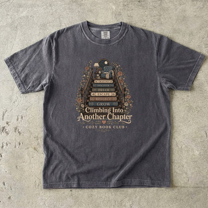 Front view of a Pepper Comfort Colors® book lover T-shirt featuring the Climbing Into Another Chapter vintage reading illustration with stacked books, flowers and a cozy reading corner.