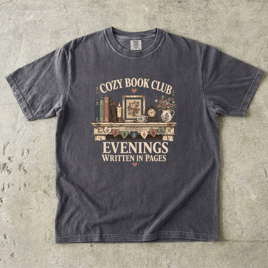 Flat lay of a Pepper Comfort Colors® book club T-shirt featuring the Cozy Book Club Evenings Written in Pages vintage bookshelf, candle, teacup and floral illustration.