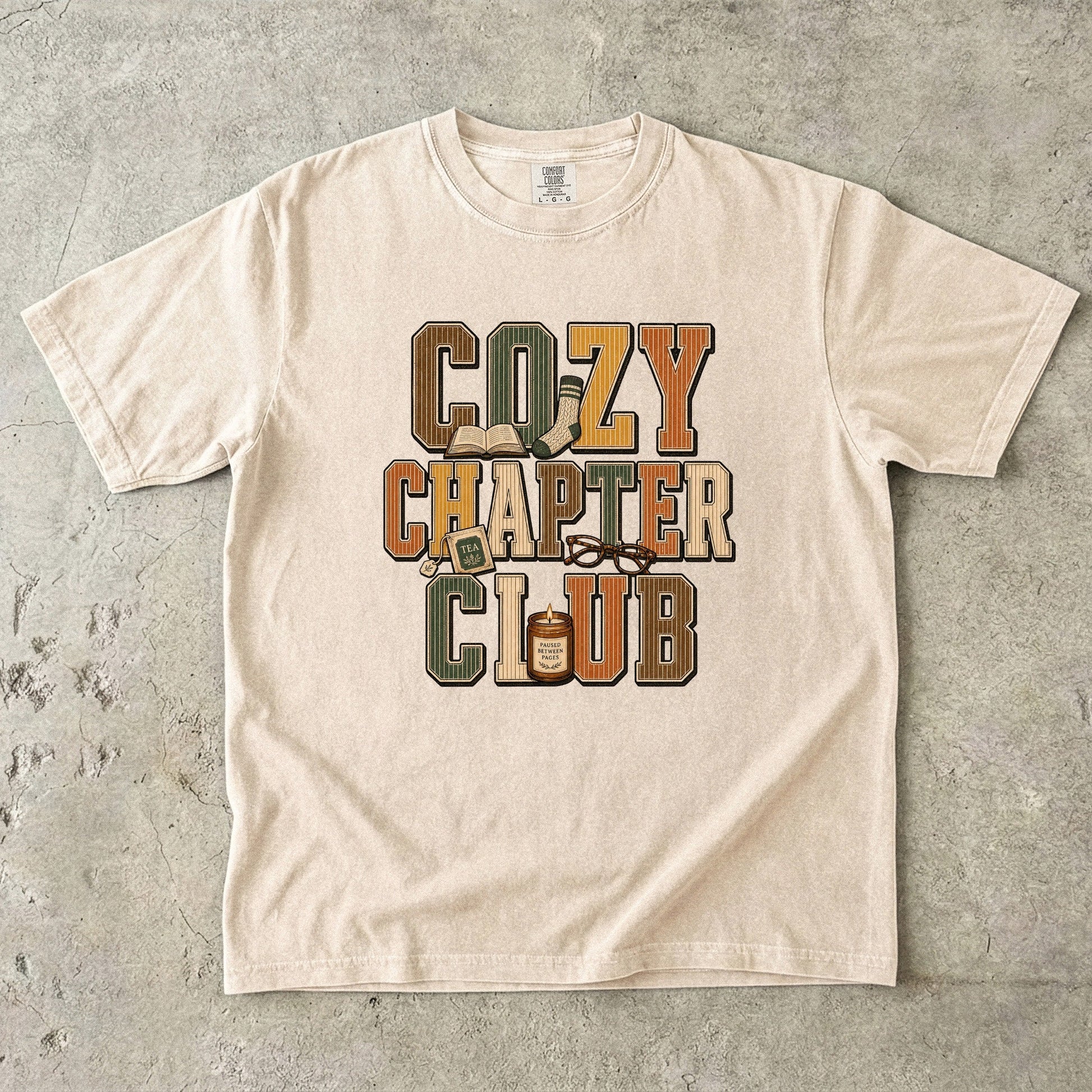 Ivory Comfort Colors Cozy Chapter Club reading T-shirt featuring retro lettering with books, tea, glasses and a candle.