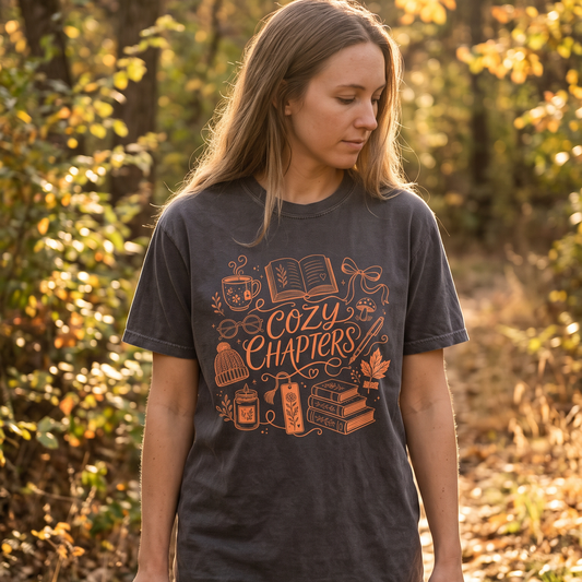 A woman standing in a sunlit autumn forest wearing a dark charcoal graphic T-shirt with orange text reading 'Cozy Chapters' and illustrated book and cozy objects, paired with taupe pants.
