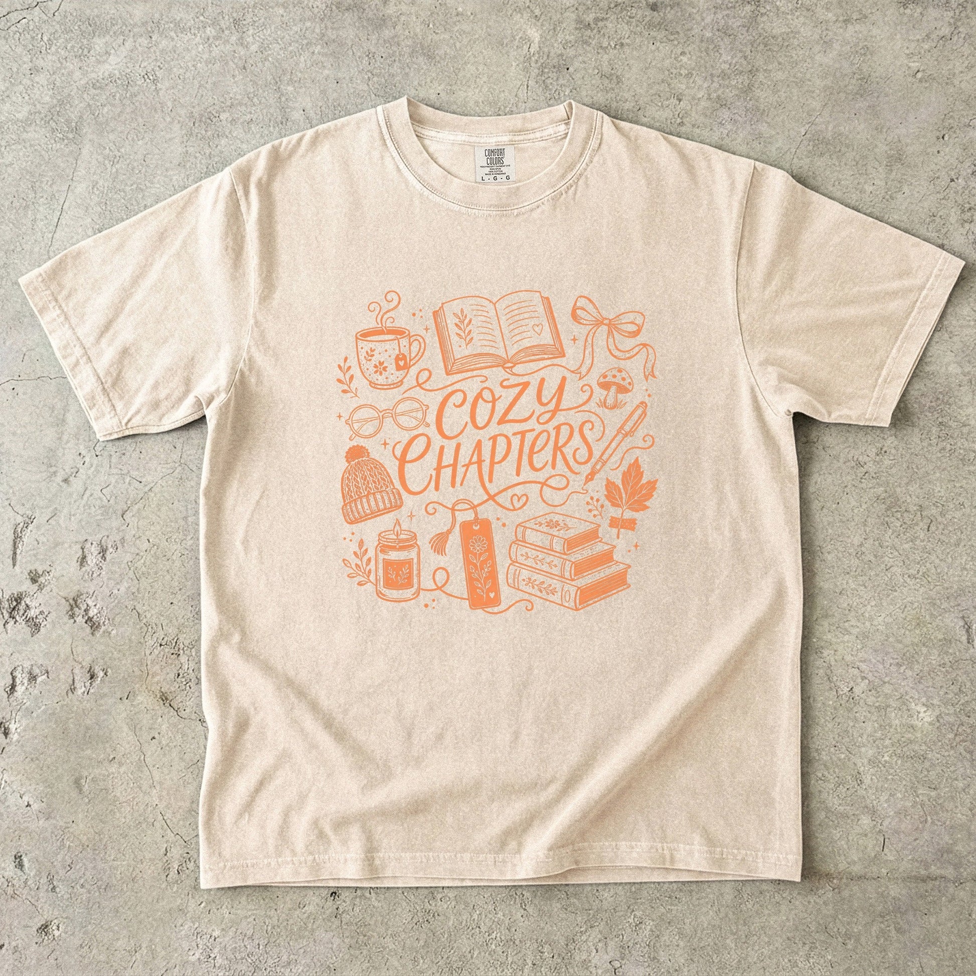 Ivory Comfort Colors book lover shirt featuring orange Cozy Chapters artwork with an open book, tea and reading accessories.