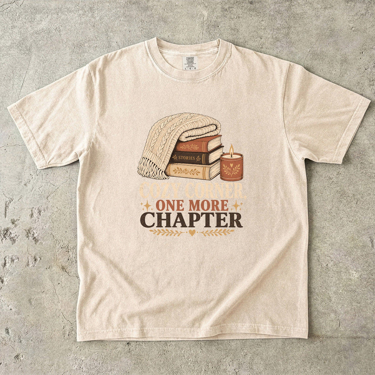 Ivory cozy reading T-shirt featuring Cozy Corner, One More Chapter with stacked books, a cable-knit blanket and glowing candle.