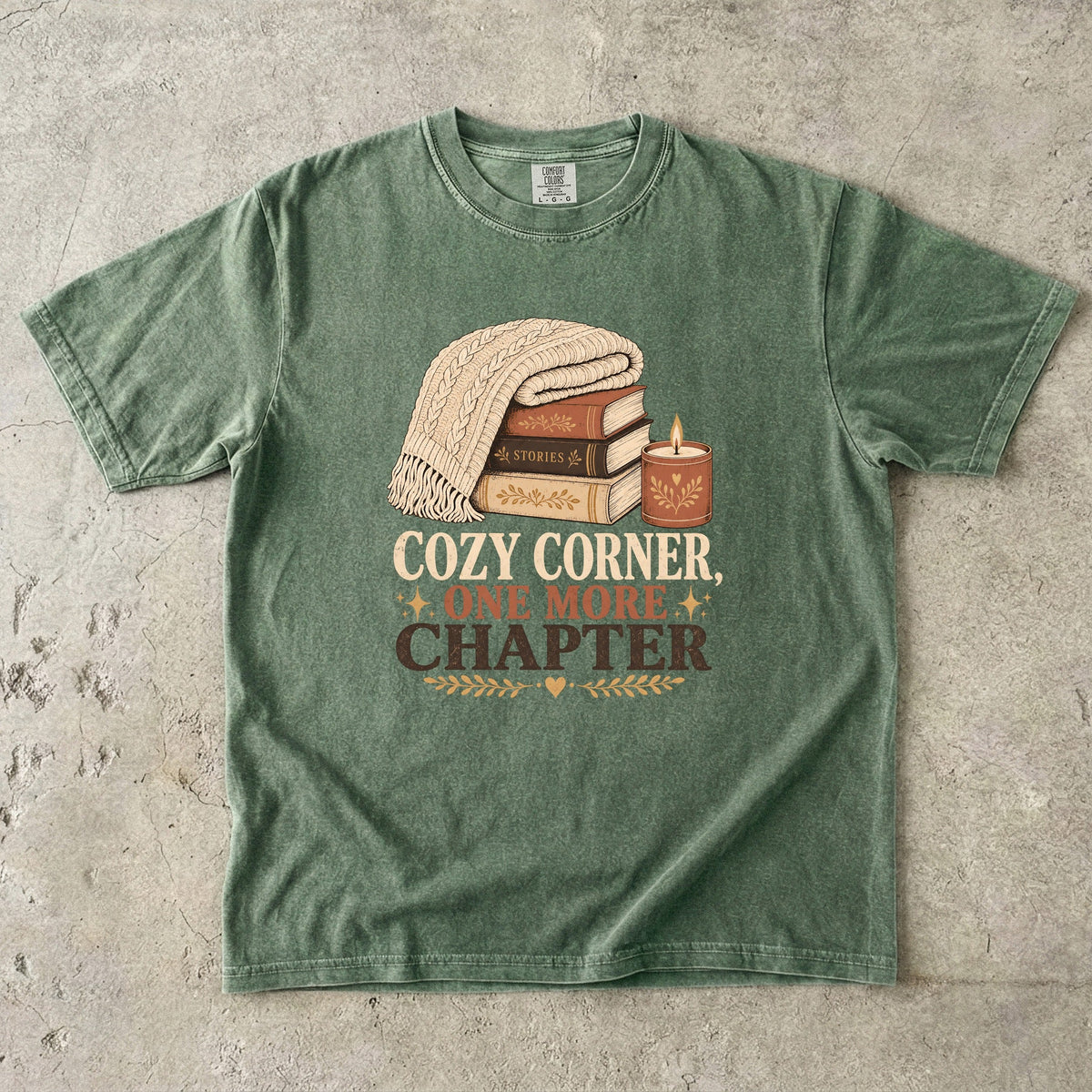 Moss cozy reading T-shirt featuring Cozy Corner, One More Chapter with stacked books, a cable-knit blanket and glowing candle.