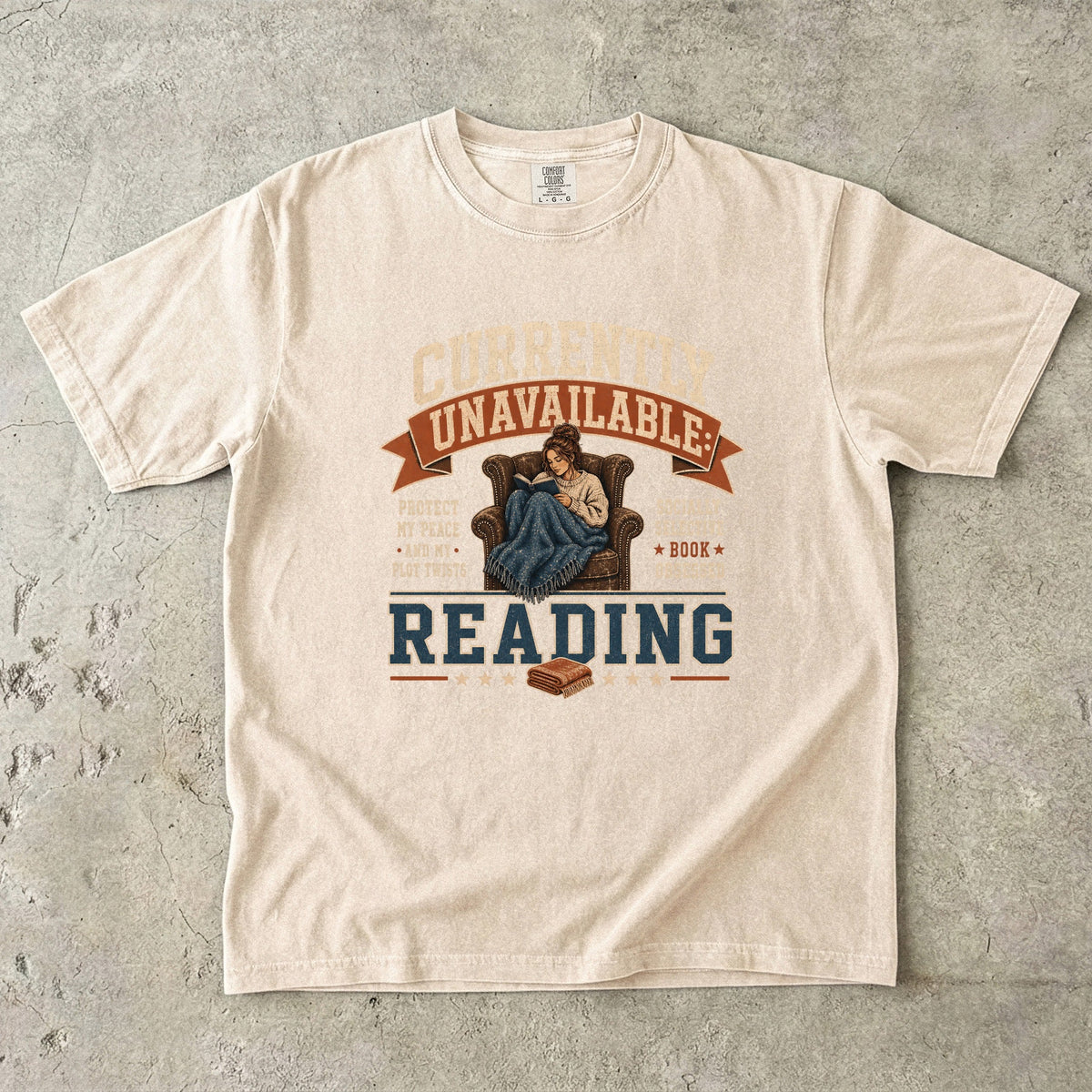 Ivory reading T-shirt featuring Currently Unavailable: Reading with a reader curled in a leather armchair beneath a blanket with a book.