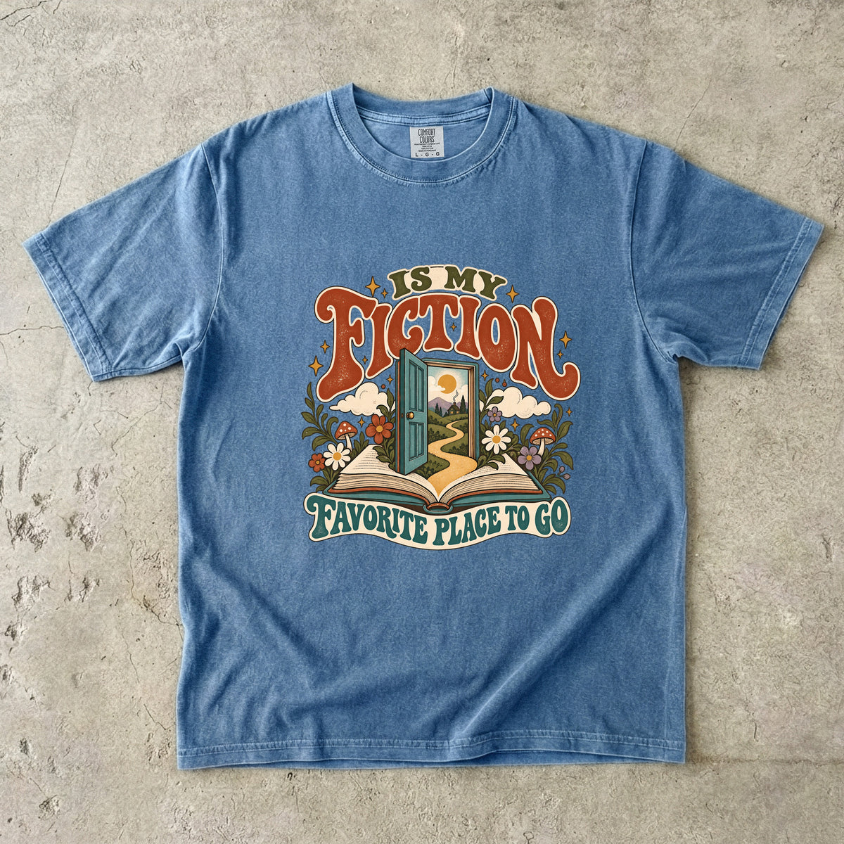 Blue Jean T-shirt featuring “FICTION / IS MY / FAVORITE PLACE TO GO” with an open book, a teal doorway leading to a winding path and cottage, flowers, mushrooms and stars.