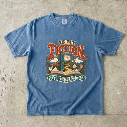Blue Jean T-shirt featuring “FICTION / IS MY / FAVORITE PLACE TO GO” with an open book, a teal doorway leading to a winding path and cottage, flowers, mushrooms and stars.