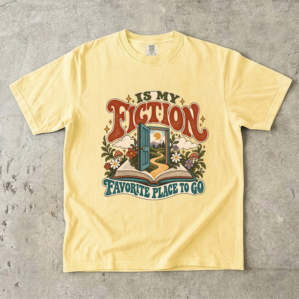 Butter T-shirt featuring “FICTION / IS MY / FAVORITE PLACE TO GO” with an open book, a teal doorway leading to a winding path and cottage, flowers, mushrooms and stars.