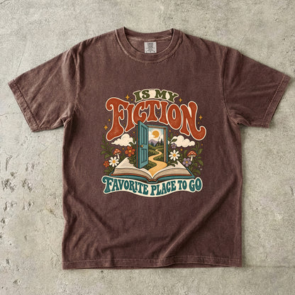 Espresso T-shirt featuring “FICTION / IS MY / FAVORITE PLACE TO GO” with an open book, a teal doorway leading to a winding path and cottage, flowers, mushrooms and stars.