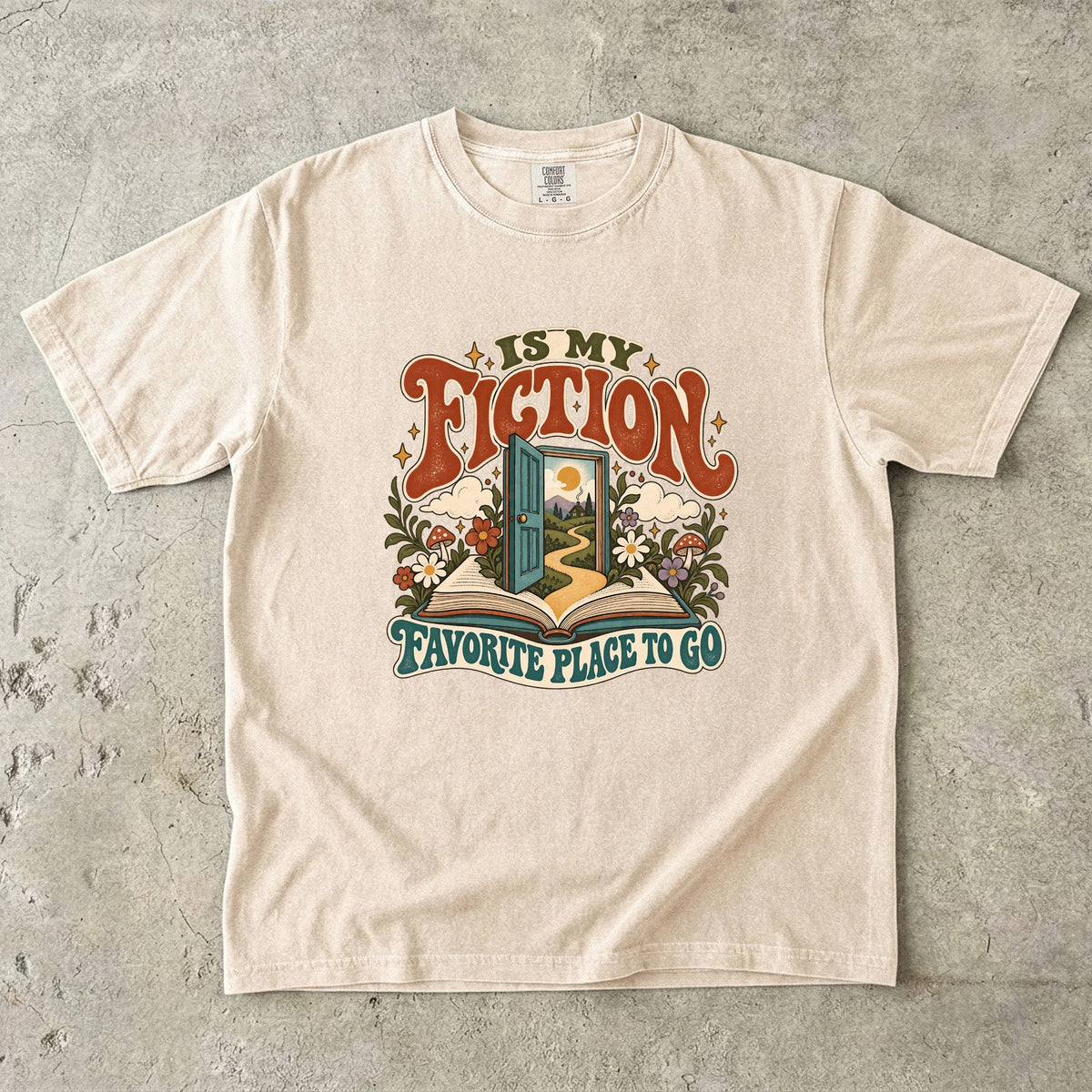 Ivory T-shirt featuring “FICTION / IS MY / FAVORITE PLACE TO GO” with an open book, a teal doorway leading to a winding path and cottage, flowers, mushrooms and stars.