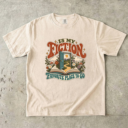Ivory T-shirt featuring “FICTION / IS MY / FAVORITE PLACE TO GO” with an open book, a teal doorway leading to a winding path and cottage, flowers, mushrooms and stars.