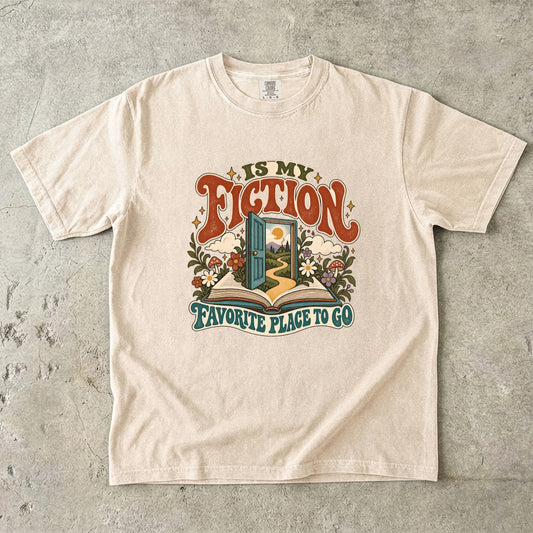 Ivory T-shirt featuring “FICTION / IS MY / FAVORITE PLACE TO GO” with an open book, a teal doorway leading to a winding path and cottage, flowers, mushrooms and stars.
