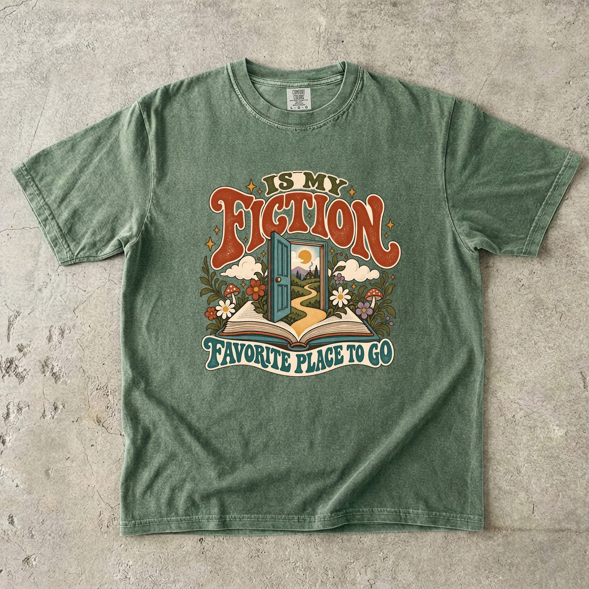 Moss T-shirt featuring “FICTION / IS MY / FAVORITE PLACE TO GO” with an open book, a teal doorway leading to a winding path and cottage, flowers, mushrooms and stars.