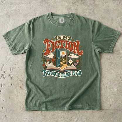 Moss T-shirt featuring “FICTION / IS MY / FAVORITE PLACE TO GO” with an open book, a teal doorway leading to a winding path and cottage, flowers, mushrooms and stars.