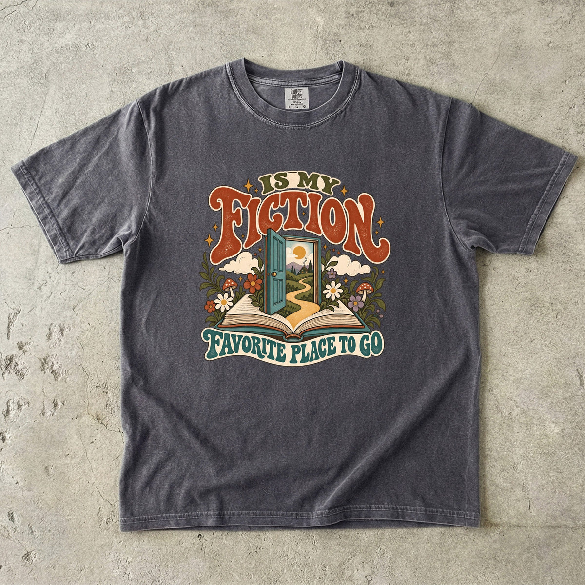 Pepper T-shirt featuring “FICTION / IS MY / FAVORITE PLACE TO GO” with an open book, a teal doorway leading to a winding path and cottage, flowers, mushrooms and stars.