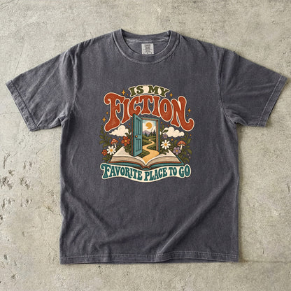 Pepper T-shirt featuring “FICTION / IS MY / FAVORITE PLACE TO GO” with an open book, a teal doorway leading to a winding path and cottage, flowers, mushrooms and stars.