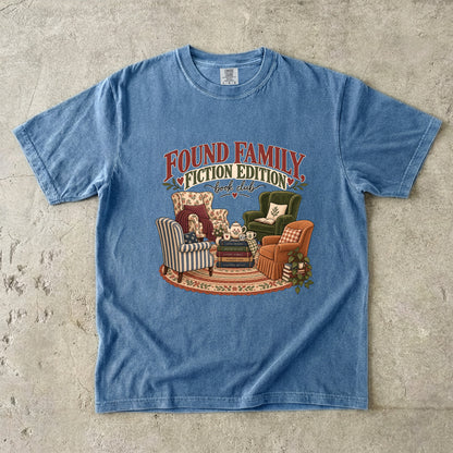 Blue Jean T-shirt featuring “Found Family, Fiction Edition” with four mismatched armchairs, books, a tea setting, cushions, a rug and leafy plants.