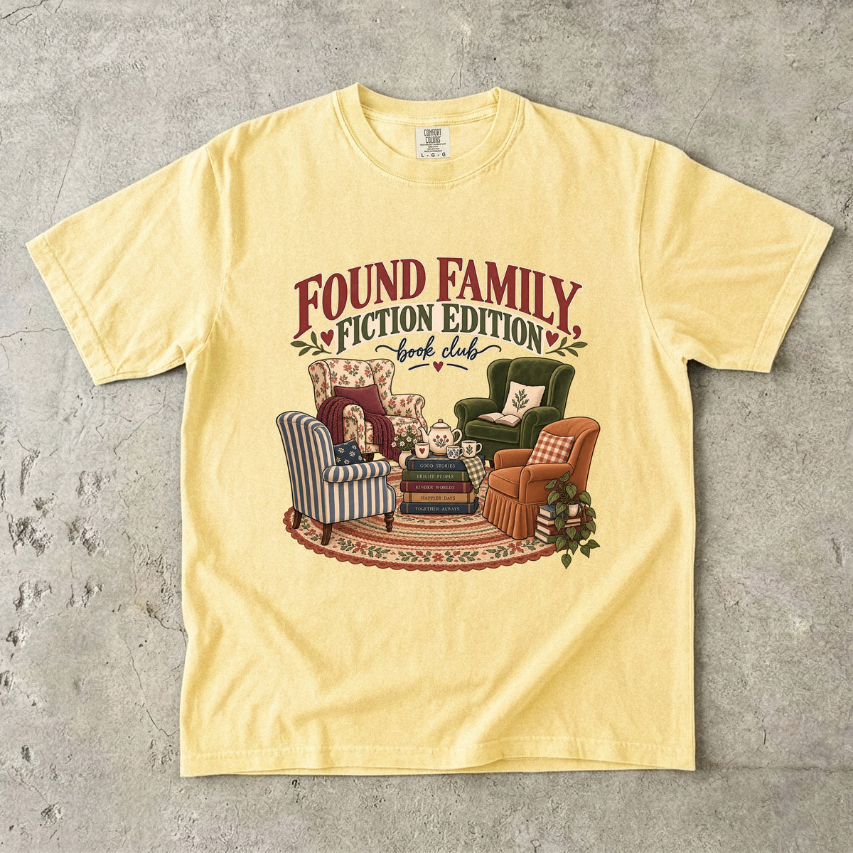 Butter T-shirt featuring “Found Family, Fiction Edition” with four mismatched armchairs, books, a tea setting, cushions, a rug and leafy plants.