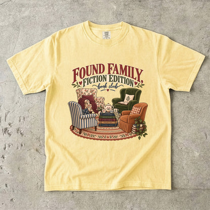 Butter T-shirt featuring “Found Family, Fiction Edition” with four mismatched armchairs, books, a tea setting, cushions, a rug and leafy plants.