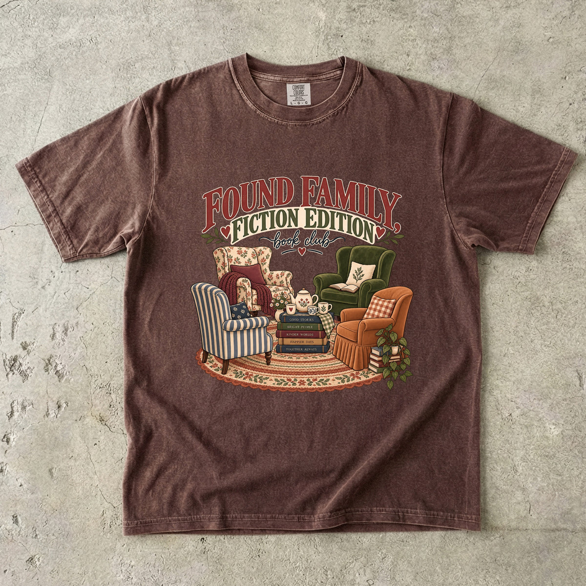 Espresso T-shirt featuring “Found Family, Fiction Edition” with four mismatched armchairs, books, a tea setting, cushions, a rug and leafy plants.