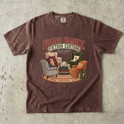 Espresso T-shirt featuring “Found Family, Fiction Edition” with four mismatched armchairs, books, a tea setting, cushions, a rug and leafy plants.