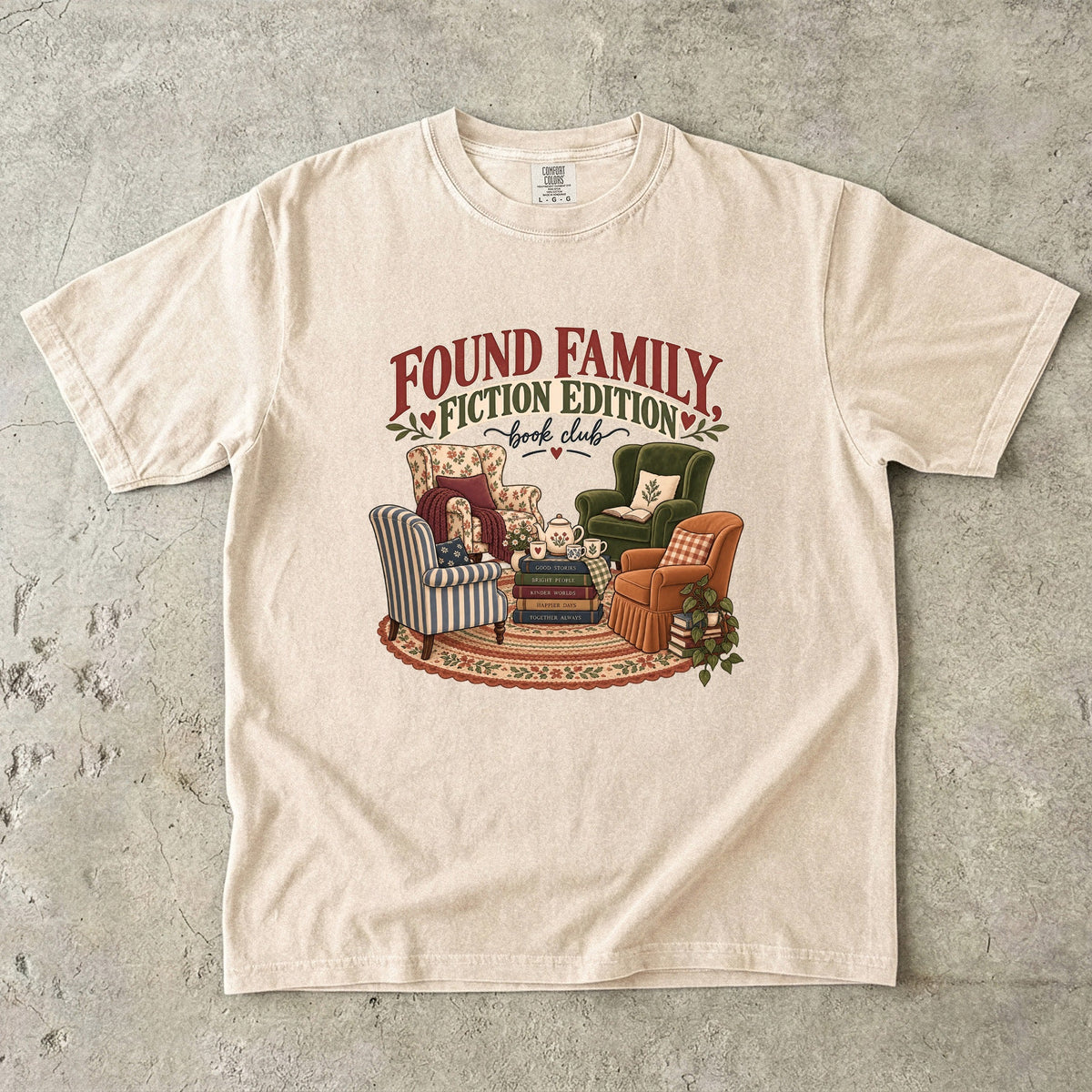 Ivory T-shirt featuring “Found Family, Fiction Edition” with four mismatched armchairs, books, a tea setting, cushions, a rug and leafy plants.