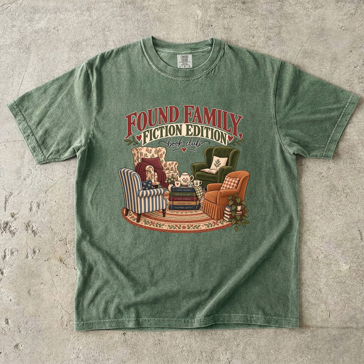 Moss T-shirt featuring “Found Family, Fiction Edition” with four mismatched armchairs, books, a tea setting, cushions, a rug and leafy plants.