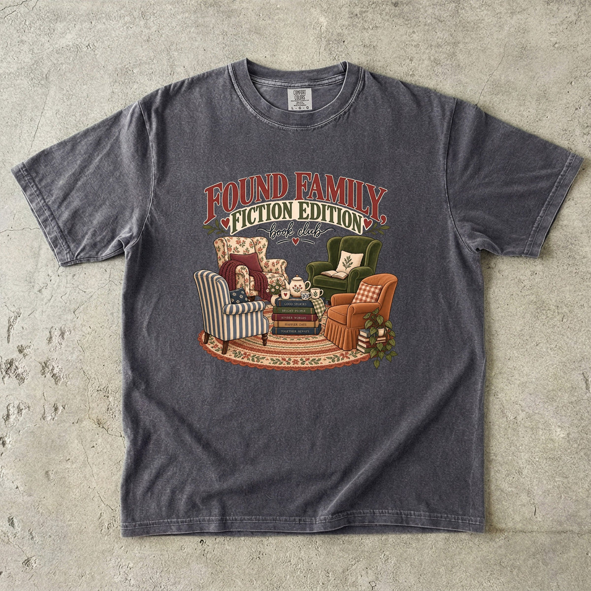 Pepper T-shirt featuring “Found Family, Fiction Edition” with four mismatched armchairs, books, a tea setting, cushions, a rug and leafy plants.